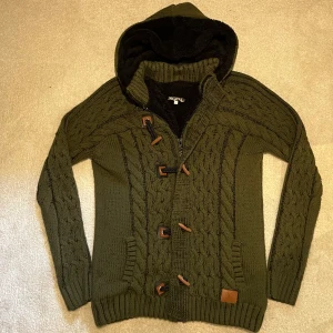 Fodrad Cardigan - Säljer en olivgrön fodrad cardigan med mysigt svart fleecefoder. Tröjan har kabelstickat mönster, huva, dragkedja och dekorativa träknappar framtill. Perfekt för kyliga sommarkvällar och riktigt skön att ha på sig. Vid minsta fråga eller fundering hör av er🤝