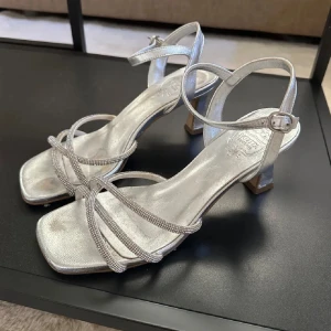 Silvriga sandaler med klack och remmar - Säljer ett par snygga silvriga sandaler med klack och tunna remmar över foten. Skorna har fyrkantig tå, justerbart spänne vid ankeln och en elegant design som passar perfekt till festliga tillfällen.