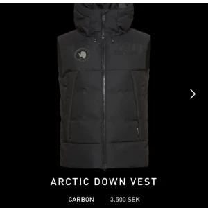 Arctic Down Vest i svart - Snygg svart dunväst med huva och dragkedja framtill. Västen har en patch på bröstet och två fickor med dragkedja. Perfekt för kyliga dagar när du vill hålla värmen utan att kompromissa med stilen.