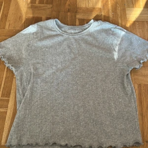 Grå ribbad t-shirt från Primark - Grå ribbad t-shirt från Primark med korta ärmar och vågiga kanter vid ärmslut och nederkant. Enkel och stilren modell som passar perfekt till jeans eller kjol. Mjuk och bekväm att ha på sig.