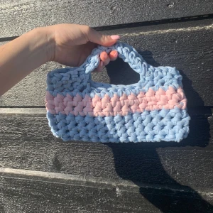 Handgjord virkad väska i blått och rosa - Superfin handgjord väska i virkad stil med breda band i ljusblått och en rosa rand i mitten. Perfekt storlek för det viktigaste och ger en söt touch till din outfit. Väskan har ett mjukt och chunky utseende. Väskan är 24cm bred och 12 cm hög