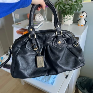Svart axelväska från Marc by Marc Jacobs - Jag säljer en svart axelväska från Marc by Marc Jacobs i skinn med guldfärgade metalldetaljer. Väskan har dubbla handtag, och insidan är fodrad med svartvitt mönster med märkets logga! I mycket fint skick, skriv vid frågor💕