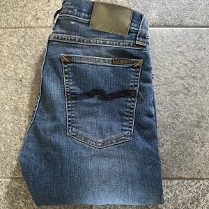 Blå jeans från Nudie Jeans - Säljer ett par klassiska blå jeans från Nudie Jeans med snygga kontrastsömmar och ikonisk broderad bakficka. Modellen har normal passform och raka ben. Perfekta till vardags för en avslappnad stil. Hör av dig vid intresse!