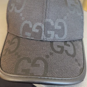 Svart keps från Gucci med GG-mönster - Säljer en svart keps från Gucci med det klassiska GG-mönstret i mörkgrått. Kepsen har böjd skärm och justerbar rem bak. Tillverkad i Italien och har en exklusiv känsla med diskret logga. Den är äkta!