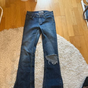 Blå lågmidjade bootcut jeans med hål på knät - Lågmidjade jeans. Säljer ett par blå bootcut jeans med klassisk femficksdesign och ett  hål på höger knä. Jeansen har normal passform och är tillverkade i denim. Perfekta för dig som gillar en avslappnad och trendig stil.