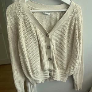 Beige stickad kofta med knappar - Säljer en beige stickad kofta med v-ringning och stora knappar framtill. Koftan har långa ärmar och bred ribbad mudd vid ärmslut och nederkant. Perfekt för lager-på-lager och enkel att matcha med olika outfits.