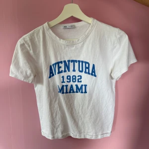 Vit t-shirt med blå text från Zara - Vit t-shirt från Zara med blå text 'Aventura 1982 Miami' på bröstet. Modellen är croppad och har korta ärmar. Perfekt för en avslappnad och cool stil. Är mer lik S i storlek!