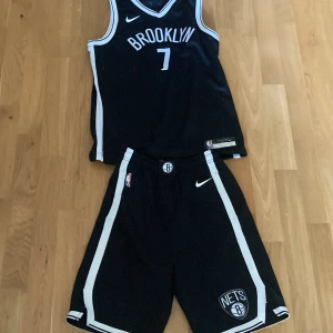 Brooklyn Nets baskettröja och shorts - Säljer en svart Brooklyn Nets baskettröja med nummer 7 och texten 'Durant' på ryggen. Tröjan är ärmlös och har vita detaljer längs sidorna. Matchande shorts med Nets-logga och vita ränder. Materialet är Dri-FIT för bra andningsförmåga.