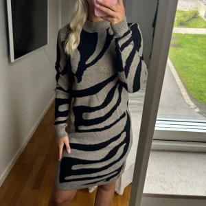 Stickad klänning med zebramönster - Snygg stickad klänning i beige och svart med zebramönster. Klänningen har lång ärm, rund halsringning och en kroppsnära passform. 
