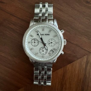 Silverfärgad klocka från Michael Kors - Stilren klocka från Michael Kors i silverfärgat rostfritt stål med länkarmband. Urtavlan är vit med glittriga stenar som timmarkeringar och har tre mindre urtavlor samt datumvisning. Perfekt accessoar för att lyfta din stil. Batteri är precis bytt och är vattentät upp till 10ATM. 