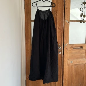 Svart långklänning, strandklänning  - Svart långklänning med linneband och lös passform. Klänningen har en enkel och stilren design med tunna axelband och lätt, luftig känsla. Perfekt för varma dagar eller som ett statement-plagg.