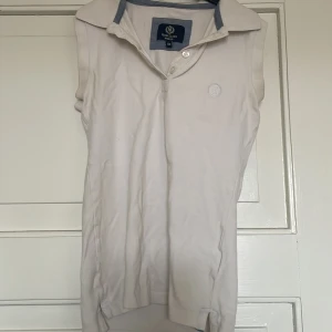 Vit ärmlös pikétopp från Henri Lloyd - Säljer en klassisk vit ärmlös pikétopp från Henri Lloyd med broderad logga på bröstet och krage med blå detalj. Toppen har knappar framtill och är tillverkad i bomull, perfekt för en sportig och stilren look.