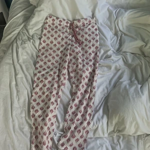 Vita byxor med rött mönster från Calliope - Säljer ett par vita byxor från Calliope med rött blommigt mönster och snörning i midjan. Byxorna har en avslappnad passform och är perfekta för varma dagar. Lätta och bekväma att bära.