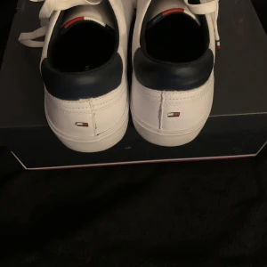 Vita sneakers från Tommy Hilfiger - Snygga vita sneakers från Tommy Hilfiger med marinblå detaljer och klassisk logga på hälen. Skorna har snörning och en platt sula. Perfekta för en stilren och sportig look.