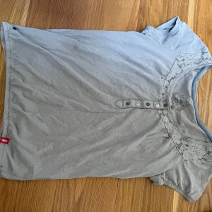 Ljusgrå topp med spetsdetaljer från edc - Säljer en ljusgrå topp från edc i storlek XS. Toppen har korta ärmar, dekorativa knappar framtill och fina spetsdetaljer längs halsringningen