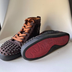 Bruna sneakers med nitar från Christian Louboutin - Säljer ett par bruna sneakers från Christian Louboutin med orange snörning och ikonisk röd sula. Skorna är täckta med svarta och silvriga nitar och har en hög modell. 