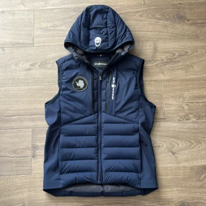 Marinblå dunväst från Sail Racing - Säljer en marinblå dunväst från Saum racing. Västen har två bröstfickor med dragkedja och en rund patch på bröstet. Perfekt för kyliga dagar och snygg till många outfits. Använd några gånger men väldigt bra skick, pris kan diskuteras