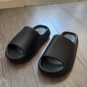 Svarta yezzy slides Onyx  - Säljer ett par svarta yezzy slides helt nya och oanvändbara . De har en minimalistisk design och är tillverkade i syntetmaterial, perfekta för sommaren eller att chilla hemma.