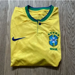 Brasilien Nike fotbollströja gul - Gul Brasilien fotbollströja från Nike med blått Swoosh och CBF-emblem broderat på bröstet. Klassisk grön och blå krage med knapp framtill. Tillverkad i Dri-FIT-material som andas, perfekt för match eller träning. Ikonisk design med detaljer i grönt och blått.