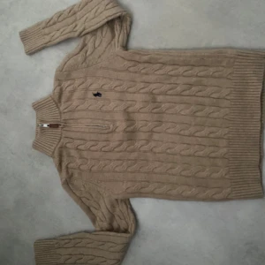 Beige stickad tröja Polo Ralph Lauren - Kabelstickad beige tröja från Polo Ralph Lauren med hög krage och dragkedja framtill. Klassisk broderad logga på bröstet och ribbade muddar. Perfekt för lager-på-lager och chill höst- eller vinterstil.