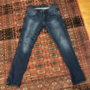 Replay blå jeans slim fit - Snygga blå jeans från Replay med klassisk femficksdesign och diskreta slitningar framtill. Jeansen har en slim passform och raka ben, perfekt för en clean och modern look. Mörkblå tvätt med ljusare partier och detaljerade sömmar.
