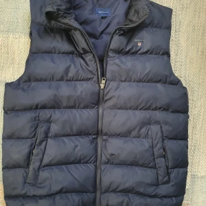 Mörkblå dunväst från GANT 158/164 - Snygg mörkblå dunväst från GANT med quiltad design och dragkedja framtill. Västen har två snedställda fickor och hög krage. Perfekt att slänga över en hoodie för extra värme. Klassisk och stilren look med GANT-logga på bröstet.