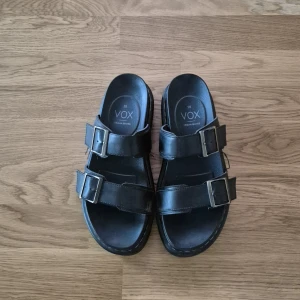 Svarta sandaler från VOX med spännen - Stilrena svarta sandaler från VOX med två breda remmar och silvriga spännen. Snygg och enkel design med platt sula, perfekt för varma dagar. Tillverkade i veganskt syntetmaterial.