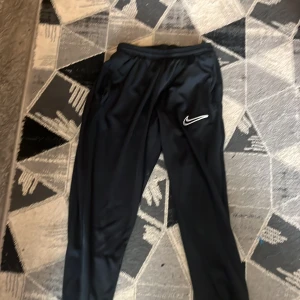 Svarta träningsbyxor från Nike - Svarta träningsbyxor från Nike med klassisk vit swoosh-logga på benet. Byxorna har elastisk midja och är gjorda i ett glansigt, lätt polyestermaterial som passar perfekt för sport eller chill. Snygg och enkel design med fickor på sidorna.