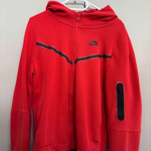 Röd Nike hoodie med dragkedja  - Nike hoodie i rött med hel dragkedja och svart logga på bröstet. Snygg svart detalj över bröstet och praktisk ficka med dragkedja på ärmen. Huva och långa ärmar gör den perfekt för kyliga dagar. Passar dig som gillar sportig och clean stil.