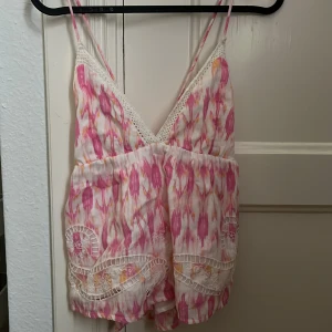 Mönstrat linne från Gina Tricot - Säljer ett superfint linne från Gina Tricot med smala axelband och v-ringning samt öppen rygg. Toppen har ett rosa och vitt mönster med inslag av orange, spetsdetaljer och broderade partier längst ner. Säljer pga att den är för stor. Inte använd.