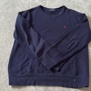 Mörkblå tröja från Polo Ralph Lauren - Snygg mörkblå långärmad tröja från Polo Ralph Lauren med klassisk rund hals och röd broderad logga på bröstet. Tillverkad i mjuk bomull.