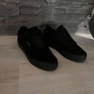 Svarta mocka sneakers från Axel Arigato - Säljer ett par svarta sneakers från Axel Arigato i mjuk mocka med diskret logga på sidan. Skorna har platt sula och rund tå, samt en stilren design utan synlig snörning. Perfekta för dig som gillar minimalistisk och clean stil. Skorna har ett litet slitage under skon men annars är dom perfekt