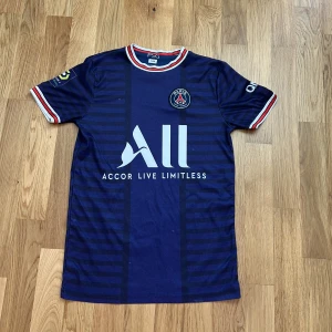 PSG Mbappé matchtröja blå 164 - Snygg PSG fotbollströja i marinblått med röda och vita detaljer på krage och ärmslut. Tröjan har Mbappé och nummer 7 på ryggen, samt klubbmärke och sponsortryck på bröstet. Tillverkad i lätt och ventilerande material, perfekt för fotbollsplanen. 