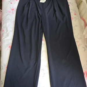 Mörkblå kostymbyxor från H&M XL - Snygga mörkblå kostymbyxor från H&M i storlek XL. Byxorna har raka, vida ben och guldiga knappar i midjan. Tillverkade i ett mjukt polyestertyg som känns bekvämt och stilrent. Perfekta för dig som gillar en klassisk och clean look.