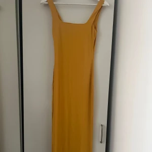 Gul långklänning med breda axelband - Säljer en snygg gul långklänning med breda axelband och rak siluett. Klänningen har en enkel och stilren design med fyrkantig urringning. Perfekt för dig som gillar minimalistisk stil och vill sticka ut med färg.