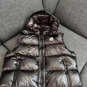 Moncler bormes väst - Säljer en svart, glansig dunväst från Moncler med huva och två dragkedjefickor framtill. Västen har klassisk Moncler-logga på bröstet och coolt serietryck på insidan. Perfekt för lager-på-lager och streetstyle.  Storlek 2 i ny skick, passar både S-M 