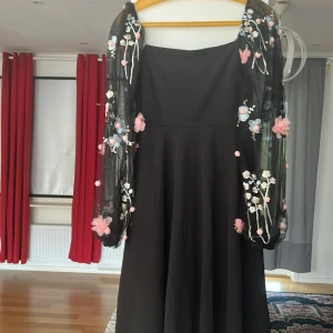 Svart klänning med broderade ärmar - Svart långklänning med transparenta långärmar i mesh, dekorerade med broderade blommor i rosa och blått samt små pärlor. Klänningen har en enkel, elegant form och är perfekt för dig som vill sticka ut med unika detaljer. Storlek 152-158 12-13 år från SHEIN 