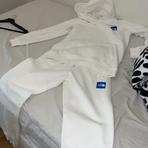 Vit tracksuit  från The North Face - Säljer en stilren vit tracksuit  från The North Face med blå logga på bröstet. Hoodien har huva, magficka och långa ärmar. Perfekt för dig som gillar en clean och enkel look. Materialet är mjuk bomull som känns skönt mot huden.