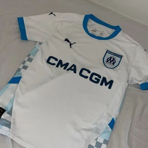 Marseille fotbollströja Puma S - Olympique de Marseille matchtröja från Puma i vitt med blå detaljer. Tröjan har klubbmärke på bröstet, sponsortryck och blå ribbad krage samt ärmslut. Sidorna har rutiga mönster i ljusblått och grått. Tillverkad i lätt funktionsmaterial.
