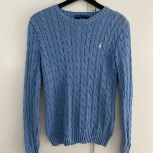 Blå kabelstickad tröja Ralph Lauren - Kabelstickad tröja från Ralph Lauren i ljusblå färg med klassisk rund halsringning och broderad vit logga på bröstet. Tröjan är långärmad och har ribbade muddar vid ärmslut och nederkant. Perfekt för lager-på-lager och snygg till jeans. Med ett en liten missfärgning på venstra axel 