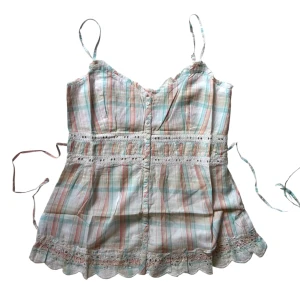 Pastellrutig linnetopp med spets - Supersöt linnetopp i pastelliga rutor med tunna axelband, knappar framtill och broderad spetskant nertill. Toppen har även spetsdetaljer vid midjan och en lätt v-ringning. Har ett band i midjan man knyter i ryggen