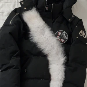 Moose Knuckles dunjacka med päls - Säljer en svart dunjacka från Moose Knuckles med vit pälsdetalj på huvan, coola metalldetaljer på ärmen och patch med hockeymotiv. Jackan har dragkedja och knappar, samt en snygg, puffig siluett som håller dig varm hela vintern.