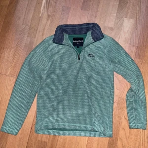 Grön fleece halfzip - Säljer en grön fleece-tröja från Weird Fish i storlek S. Tröjan har ett diskret rutmönster, halv dragkedja framtill och en mörkgrå krage med struktur. Broderad logga på bröstet och mjukt material som är perfekt när det är lite kyligt.