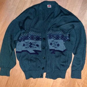 Mörkgrön cardigan - Säljer en mörkgrön stickad kofta från NG i storlek M. Koftan har ett snyggt geometriskt mönster i grått och mörkblått över bröstet, knappar framtill och ribbade muddar vid ärmar och nederkant. Perfekt för dig som gillar retrovibbar och vill ha något unikt i garderoben.
