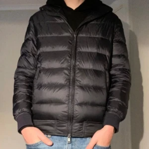 Svart dunjacka - Säljer en svart pufferjacka med huva och dragkedja framtill. Jackan har ett quiltat mönster och ribbade muddar vid ärmslut. Perfekt för kalla dagar och har en clean, modern look som funkar till jeans och hoodie. Långärmad och med praktiska fickor på sidorna.