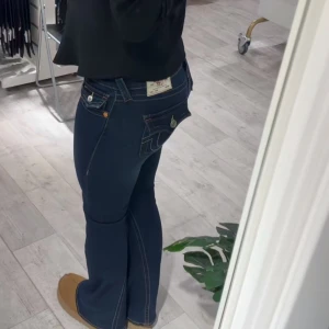Mörkblå True Religion bootcut jeans - INTRESSEKOLL⚠️Snygga mörkblå jeans från True Religion med klassiska kontrastsömmar och ikoniska fickdetaljer med silverknappar. Modellen har bootcut-ben och låg midja, vilket ger en schysst siluett. Jeansen är tillverkade i stretchigt denim för extra komfort.