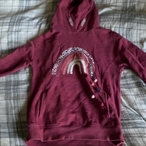 Vinröd hoodie med regnbågsmotiv från Next - Säljer en vinröd hoodie från Next med ett coolt regnbågsmotiv framtill, där regnbågen har leopardmönster och små färgglada bollar som detalj. Tröjan har huva, långa ärmar och är gjord i mjuk bomull. Perfekt för dig som gillar färg och unika prints!