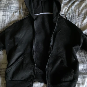 Svart hoodie med Mona Lisa-tryck från SHEIN - Svart hoodie från SHEIN i storlek XS, tillverkad i 100% polyester. På ryggen finns ett coolt tryck med Mona Lisa och en vit grafisk illustration med handtecken. Hoodien har huva, dragkedja och två fickor framtill. Perfekt för dig som gillar streetstyle och unika prints.