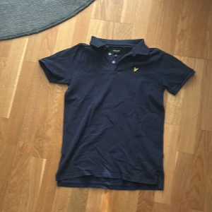 Marinblå pikétröja Lyle & Scott Junior - Snygg marinblå pikétröja från Lyle & Scott Junior med klassisk krage, korta ärmar och gul broderad logga på bröstet. Tillverkad i bomull för en skön känsla och stilren look. Perfekt för dig som gillar enkel men ikonisk design.