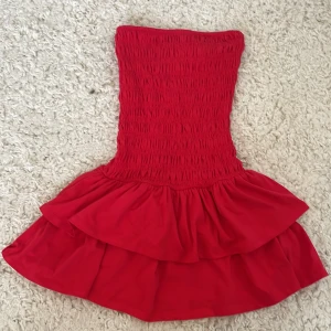 Röd volangklänning från Gina Tricot - Supersöt röd kortklänning från Gina Tricot med smockad överdel och dubbla volanger i kjolen. Axelbandslös modell som sitter snyggt och är perfekt för sommarens alla tillfällen. Materialet känns lätt och följsamt.